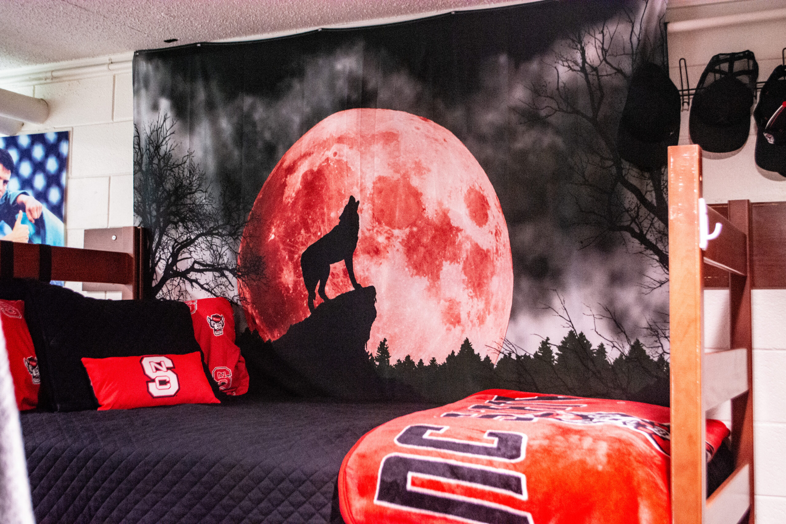 Bed with NC State pillows and blankets. Red moon with wolf silhouette tapestry.