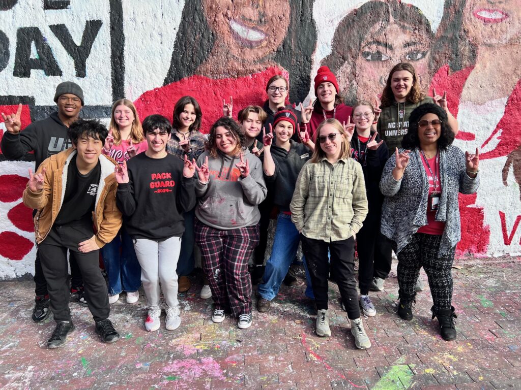 Arts village students standing in front of the mural they painted for ESPN game day holding up wolfies. 